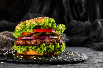 Canvas Print - Beef burger with tomatoes, red onions, cucumber and lettuce on black slate over dark background. Unhealthy food