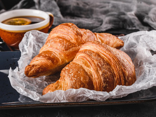 Wall Mural - Breakfast with fresh french chocolate croissants on paper over dark background with napkin and cup of tea. Dessert, puff pastries, top view