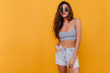 Glad shapely dark-haired girl in black sunglasses standing on yellow background. Indoor photo of slim lady in denim shorts and tank-top.