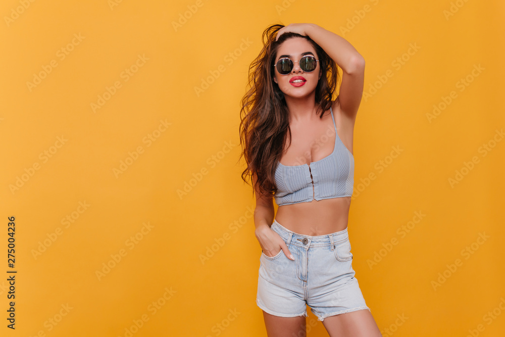 Happy girl in vintage denim shorts standing in confident pose on yellow background. Indoor portrait of enchanting young lady in summer casual outfit plays with her hair. - obrazy, fototapety, plakaty 