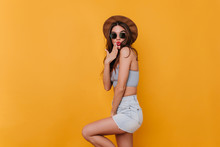 Refined Shapely Girl Wears Blue Denim Shorts Playfully Posing In Studio. Indoor Portrait Of Graceful Dark-haired Female Model In Hat And Sunglasses.