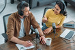 © LIGHTFIELD STUDIOS - cheerful radio host talking to colleague while sitting at wooden table near microphones in radio studio