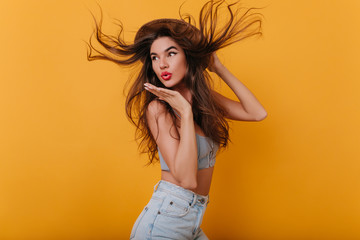 Wall Mural - Amazing girl with long straight hair dancing in studio and sending air kiss. Indoor portrait of lovely brunette female model in hat and denim pants isolated on yellow background.