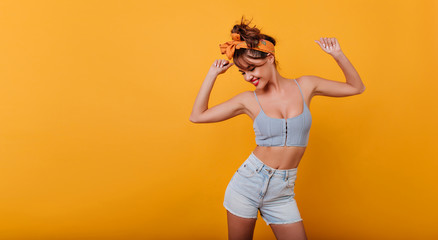 Sporty lovely girl in tank-top and denim shorts dancing with pleasure and smiling. Indoor portrait of stunning female model with vintage hairstyle waving hands on yellow background.