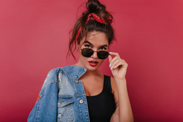 Wall Mural - Fashionable dark-haired woman in black sunglasses looking with interest on claret background. Enthusiastic girl with vintage denim jacket posing in studio.