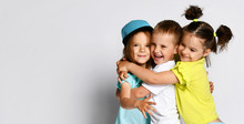 Studio Portrait Of Children On A Light Background: Full Body Shot Of Three Children In Bright Clothes, Two Girls And One Boy. Triplets, Brother And Sisters. Hugging On Camera. Family Ties, Friendship