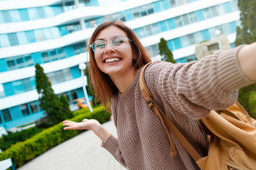 Wall Mural - Young girl take selfie from hands with phone on summer spring city street. Urban life concept.Closeup selfie-portrait student of attractive girl in sunglasses with long hairstyle and snow-white smile 