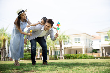 Wall Mural - Happy Asian family. Father gave his daughter a piggyback at a park at natural sunlight background and house. Family vacation concept with copy space