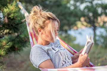 Wall Mural - young attractive caucasian woman reading book in hammock in the forest, lake on the background