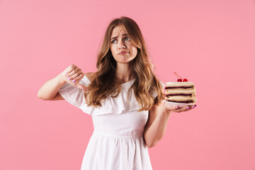 Wall Mural - Image of disappointed sad woman wearing white dress holding piece of cake with thumb down