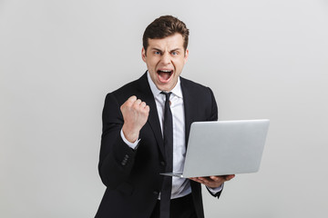 Wall Mural - Image of successful excited businessman in formal suit celebrating triumph while holding laptop isolated over gray background