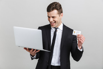 Canvas Print - Image of happy brunette businessman in formal suit laughing while holding laptop and credit card