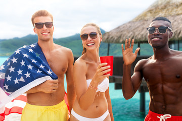 summer, holidays and people concept - group of happy friends with flag and non alcoholic drinks celebrating american independence day and party over bungalow on tropical beach in french polynesia