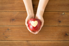 Food, Valentines Day And Health Concept - Close Up Of Hands Holding Ripe Red Apple With Carved Heart Shape Over Wooden Table