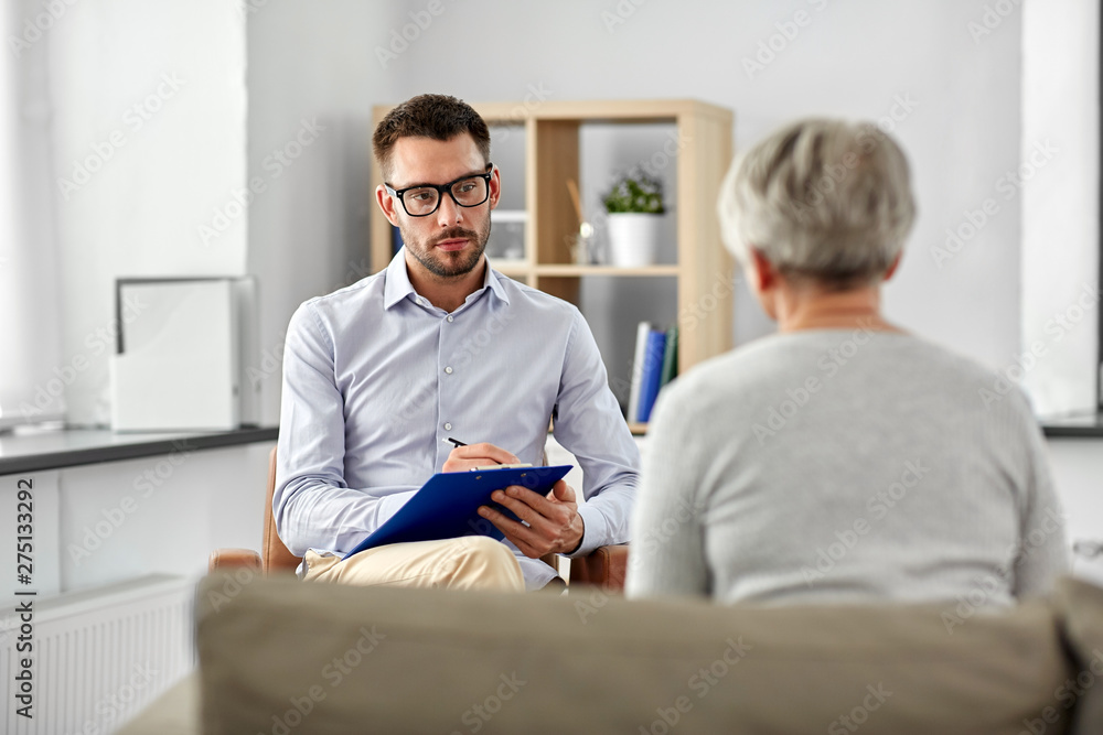 geriatric psychology, mental therapy and old age concept - psychologist with clipboard taking notes and listening to senior woman patient at psychotherapy session - obrazy, fototapety, plakaty 