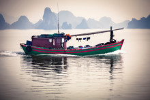 Vietnamese Junk Fishing Boat Crossing The Water In Halong Bay, Vietnam With The Famous Limestone Stacks In The Background