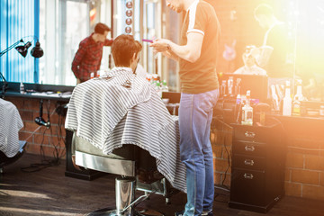 Master cuts hair and beard of men in the barbershop, hairdresser makes hairstyle for a young man. Barber work with clipper machine in barbershop.