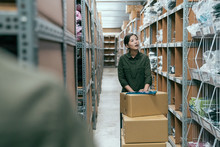 Young Asian Female Delivery Postman Pushing Cart Walking In Storehouse Of Company. Woman Moving Lots Of Cardboard Boxes While Doing Stocktaking In Warehouse. Girl Staff In Uniform Work In Stockroom
