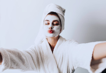 Self portrait of charming, stylish, pretty, model after bath wrapped in towel shooting selfie on front camera applying using face mask for her dry, oiled, sensual face skin, enjoying procedure