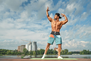 Professional bodybuilder posing on lake pier with hands up.