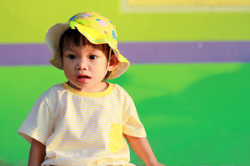 Portrait of happy Asian baby child girl. She wearing a yellow shirt and hat. Sweet smiling. On green and colorful background. Travel in the summer season concept. Baby aged 1 year and 4 months olds.