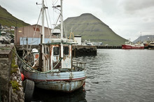 View On Industrial Harbour: Old, Rusty, Abandoned Fishing Boat Moored In Docks, Big Green Hills, Some Of Them Covered With Grey Clouds In The Background; Faroe Islands