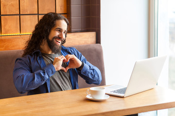 Love you! Portrait of romantik handsome young adult man freelancer in casual style sitting in cafe and talking with his friend in laptop, showing heart sign with fingers. Indoor, lifestyle concept