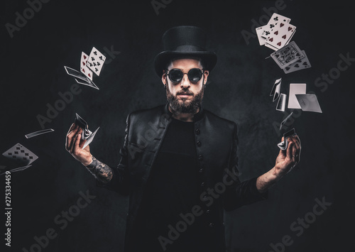 Fotomural Magician in a black suit, sunglasses and top hat, showing trick with playing cards on a dark background