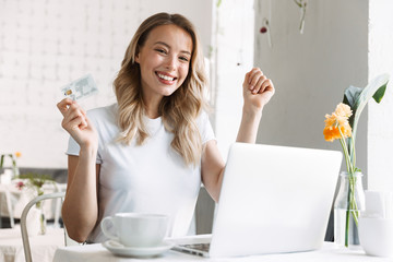 Poster - Pleased young pretty blonde woman student sitting in cafe using laptop computer drinking coffee holding credit card.