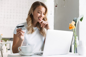 Wall Mural - Happy young pretty blonde woman student sitting in cafe using laptop computer drinking coffee holding credit card.