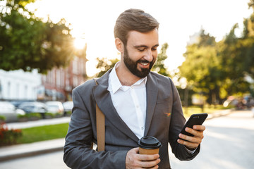 Poster - Portrait of handsome adult businessman in formal suit holding takeaway coffee and using smartphone outdoors