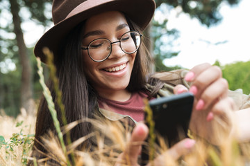 Poster - Photo of joyful stylish woman wearing hat and eyeglasses using cellphone while lying on grass in green park