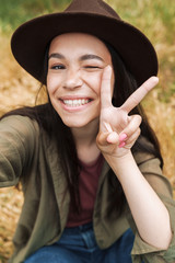 Sticker - Photo closeup of young woman wearing hat smiling and showing peace sign while taking selfie photo outdoors