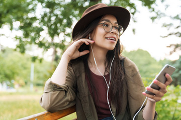 Sticker - Photo of nice woman wearing hat and eyeglasses using earphones and smartphone in green park