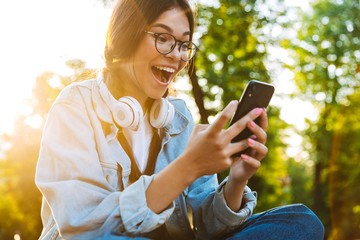 Surprised cute young student girl wearing eyeglasses sitting outdoors in nature park using mobile phone.