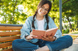 © Drobot Dean - Cheerful cute young student girl wearing eyeglasses sitting on bench outdoors in nature park writing notes in notebook.