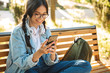 © Drobot Dean - Pleased positive cheerful cute young student girl wearing eyeglasses sitting on bench outdoors in nature park using mobile phone.