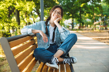 Poster - pleased happy cute young student girl wearing eyeglasses sitting on bench outdoors in nature park