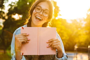 Wall Mural - Cheerful cute young student girl wearing eyeglasses sitting outdoors in nature park holding book.