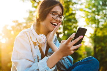 Canvas Print - Happy cute young student girl wearing eyeglasses sitting outdoors in nature park using mobile phone.