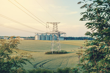 Metal support is a pillar of a high-voltage line in the field. Big elevator on the background. Industrialization of agriculture