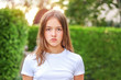 Close-up portrait of serious angry teenager girl outdoors in park with green hedge background. No makeup, natural beauty concept. Summer lifestyle. Facial expression, teenager period