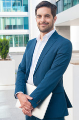 Smiling business man holding laptop and posing outdoors. Handsome young guy standing and looking at camera with blurred building in background. Successful businessman portrait concept. Front view.