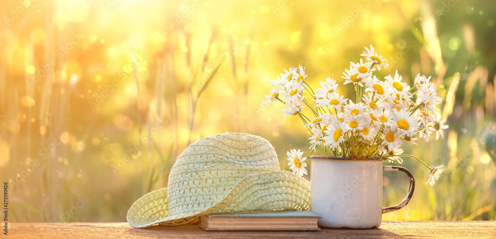 beautiful composition with chamomile flowers in Cup, old book, braided hat in summer garden. Rural landscape natural background with Chamomile in sunlight. Summertime season. copy space - obrazy, fototapety, plakaty 