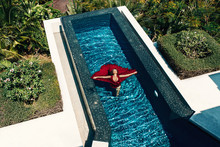 Fashion  Aerial Photo: Girl  With Bright Make Up In A Red Dress Lying On The Water Of The Pool. Young Woman With Closed Eyes Posing In Water