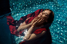 Fashion Photo: Girl  With Bright Make Up In A Red Dress Lying On The Water Of The Pool. Young Woman With Closed Eyes Posing In Water