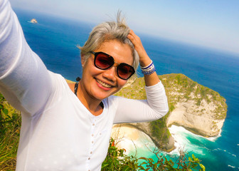 middle age 50s happy and cheerful Asian woman with grey hair taking selfie with mobile phone at beautiful tropical beach island smiling at cliff viewpoint enjoying Summer