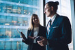© BullRun - Corporate business people communicating and sharing opinions about digital tablet while standing in office interior near big window, man and woman colleagues talking and discussing work project