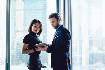 Wall Mural - Male and female lawyers checking information on touch pad standing near skyscraper window in office, successful diverse employees reading financial news on digital tablet, colleagues collaborating