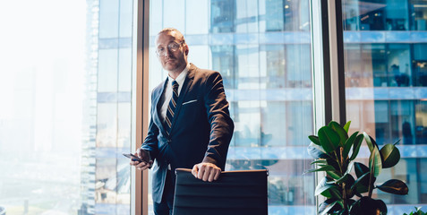 Half length portrait of intelligent male corporate manager formally dressed holding cellular phone for banking and looking at camera during working time in office, business man in spectacles